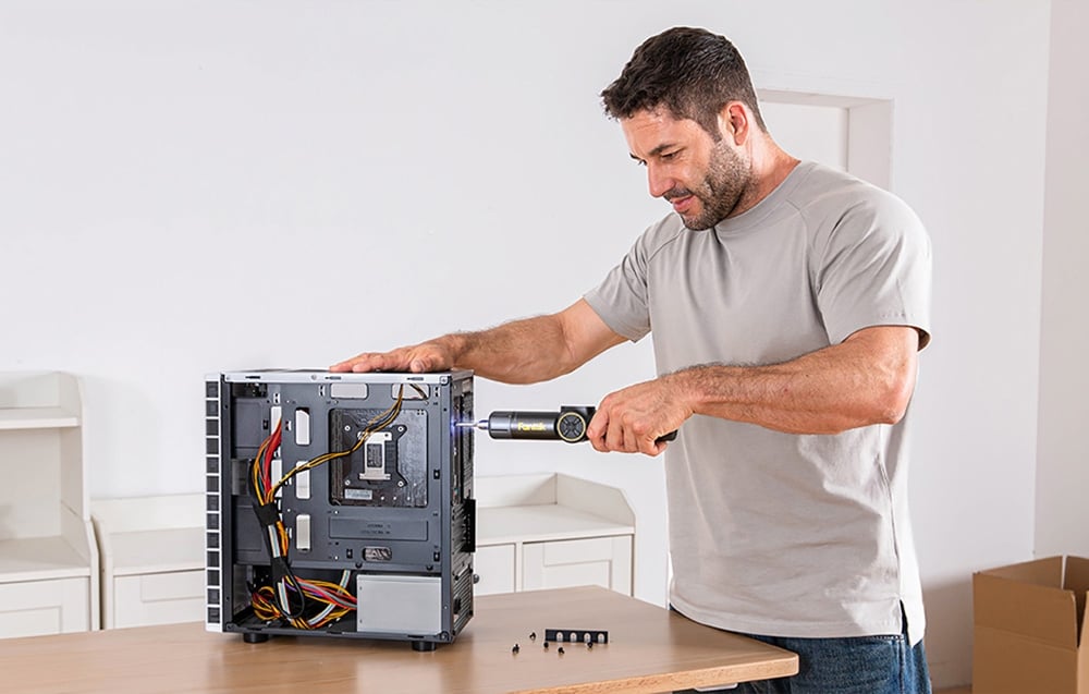Un homme en T-shirt gris et jeans répare un ordinateur avec une visseuse électrique.