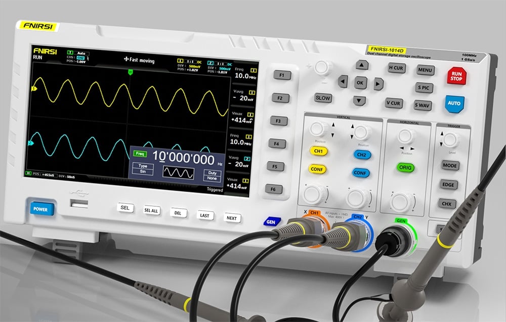 Oscyloskop na biurku z podłączonymi przewodami, na ekranie widoczne żółty i niebieski wykres sinusoidalny.