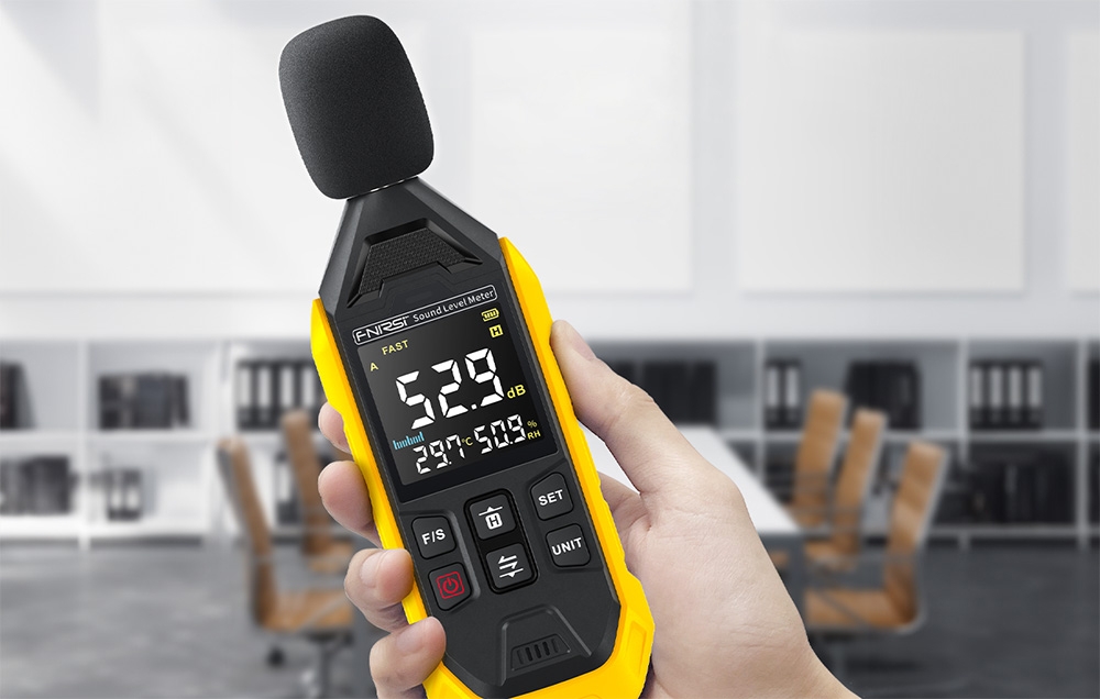 Main tenant un mètre décibel avec protection de microphone et affichage montrant 52,9 dB dans un bureau.