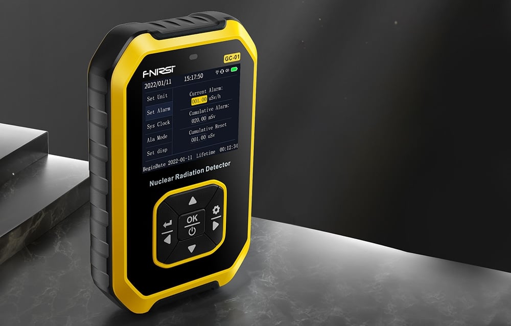 Détecteur de radiation nucléaire avec un boîtier jaune et noir sur une surface en pierre, affichant l'écran avec les paramètres d'alarme et de mesure.
