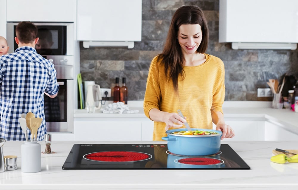 Une femme en pull jaune cuisine dans une casserole bleue sur une plaque en céramique avec des brûleurs rouges