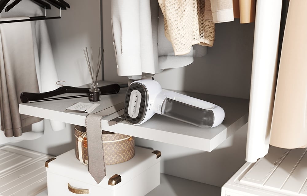 Un défroisseur blanc se tient sur une étagère blanche dans un dressing entre des vêtements suspendus et un diffuseur de parfum