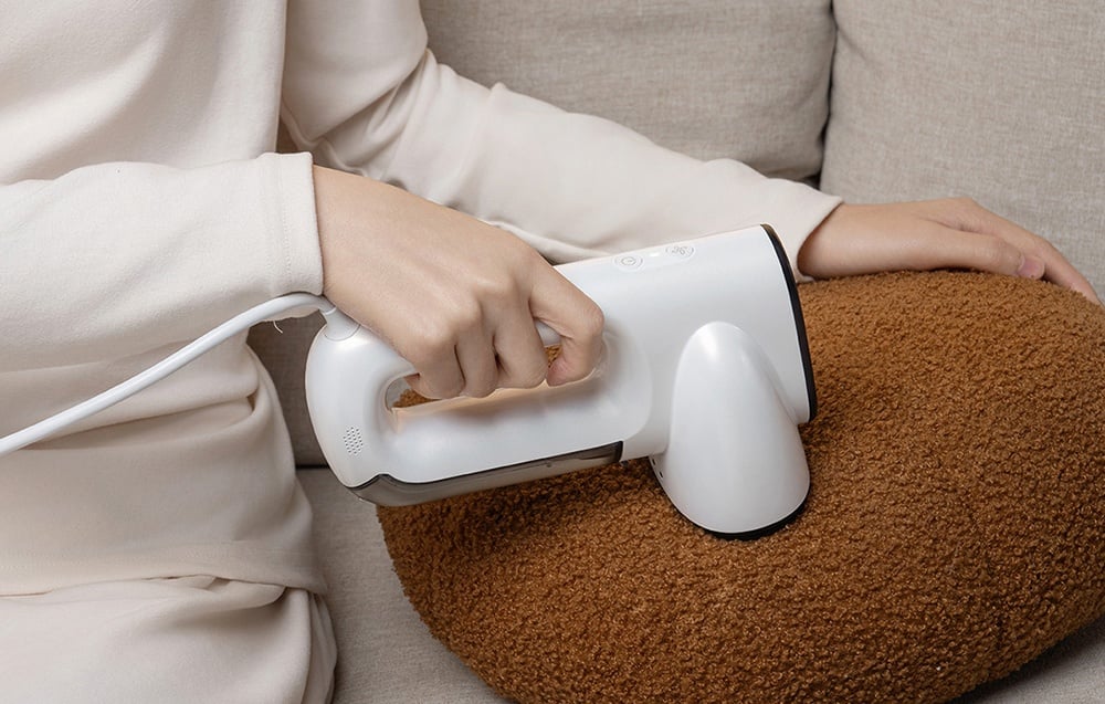Une personne en tenue beige est assise sur un canapé et utilise un défroisseur blanc pour nettoyer un coussin marron