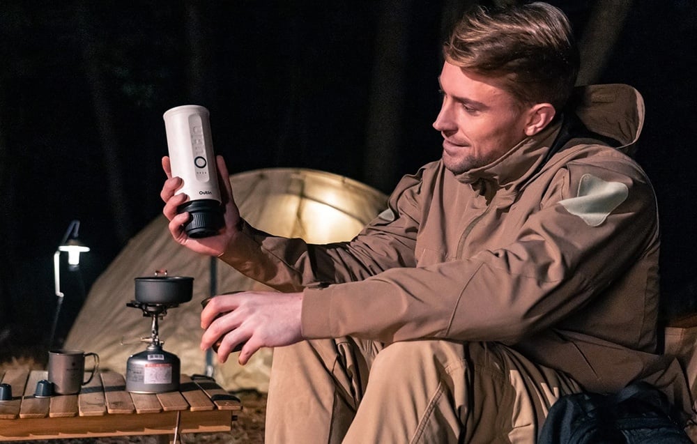 Un homme en veste marron prépare du café avec la machine à café sans fil Outin. Une tente est visible en arrière-plan.