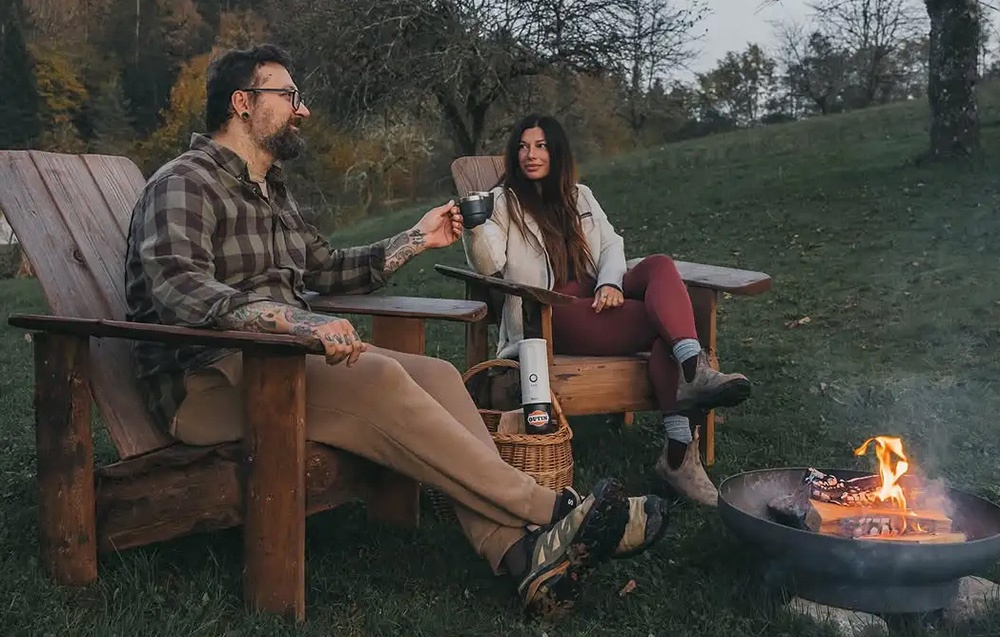 Un homme et une femme assis sur des chaises boivent du café. Un feu de camp brûle devant eux.