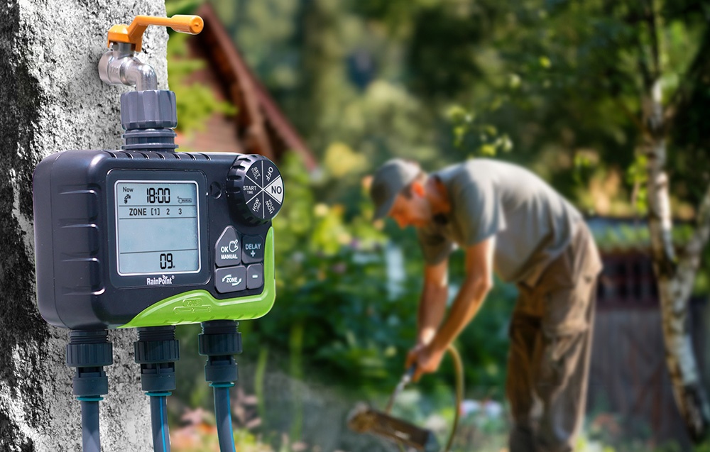 Minuteur d'arrosage RainPoint installé sur un mur de jardin, en arrière-plan une personne arrosant des plantes
