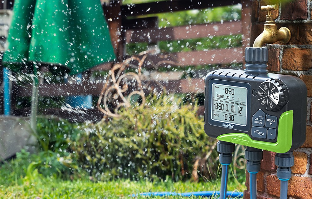 Contrôleur RainPoint connecté à un robinet sur un mur en brique, à côté d'un arroseur fonctionnant dans le jardin