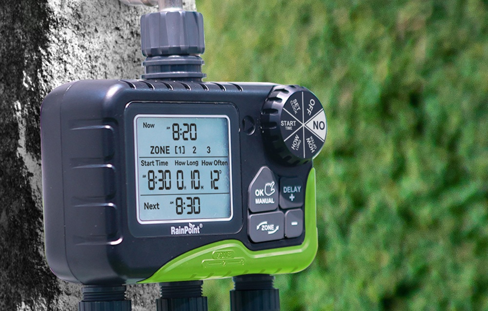 Gros plan sur le contrôleur d'arrosage RainPoint fixé à un arbre, en arrière-plan une haie verte