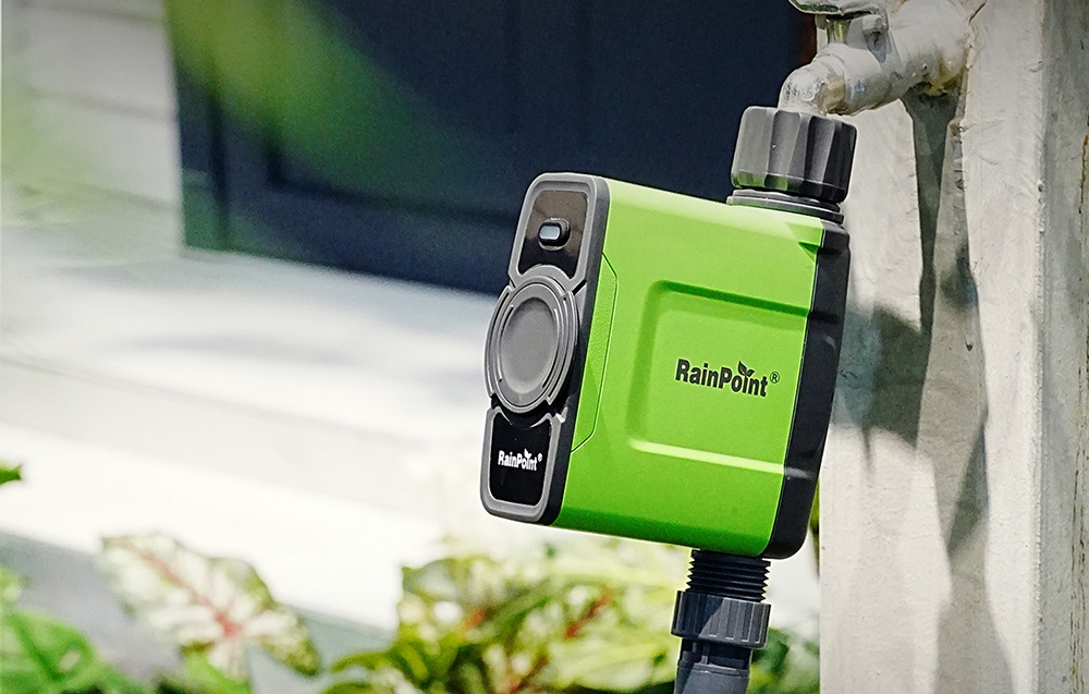 Contrôleur RainPoint fixé à un tuyau près de la maison entouré de plantes de jardin