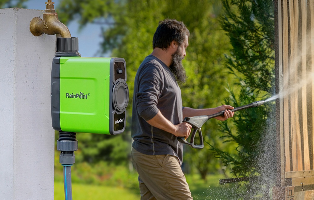 Contrôleur d'irrigation RainPoint monté sur un poteau blanc, en arrière-plan un homme nettoyant un mur en bois avec un nettoyeur haute pression